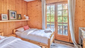 A wooden cabin room with two beds, a shelf with framed pictures, a hanging lamp, and a large window with curtains. - Le Chalet des Neiges - Welkeys (Megève)