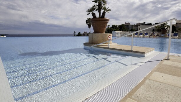 Et klart swimmingpool med fliseterrasse, plantekasse med palmetræ og et forbudsskilte mod svømning.