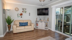 Un salon avec un canapé beige, un téléviseur à écran plat, un plancher en bois et une porte-fenêtre coulissante donnant sur l’extérieur.