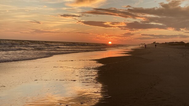Une plage au coucher du soleil, avec des vagues et quelques personnes qui se promènent.