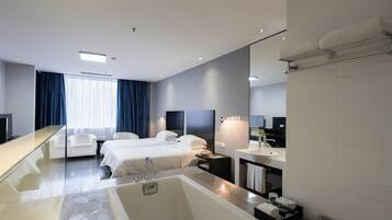 A modern hotel room with a large bathtub, two beds, a sitting area with chairs, a vanity with a mirror, and a large window with curtains.