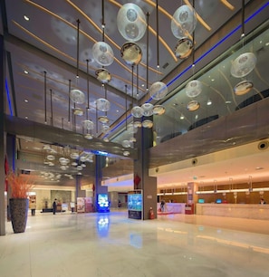A spacious lobby with a modern design, featuring a large reception desk, a digital information kiosk, and a large potted plant.