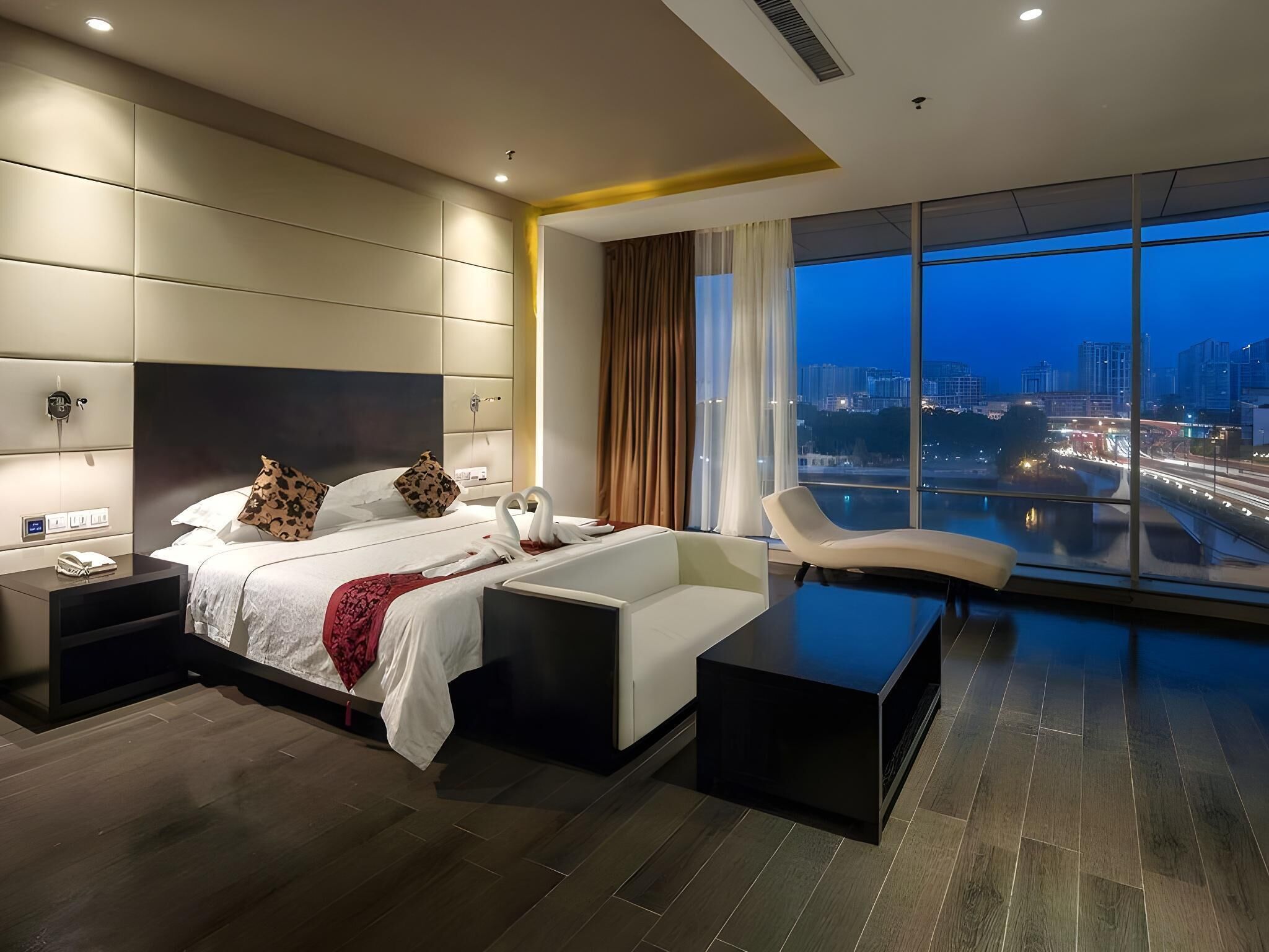 Une chambre d’hôtel moderne dotée d’un grand lit, d’un coin salon et offrant une vue sur la ville au crépuscule.
