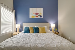 Un lit bien fait, recouvert d’une couette blanche, avec deux lampes de chevet et un mur bleu orné d’une œuvre d’art abstraite.