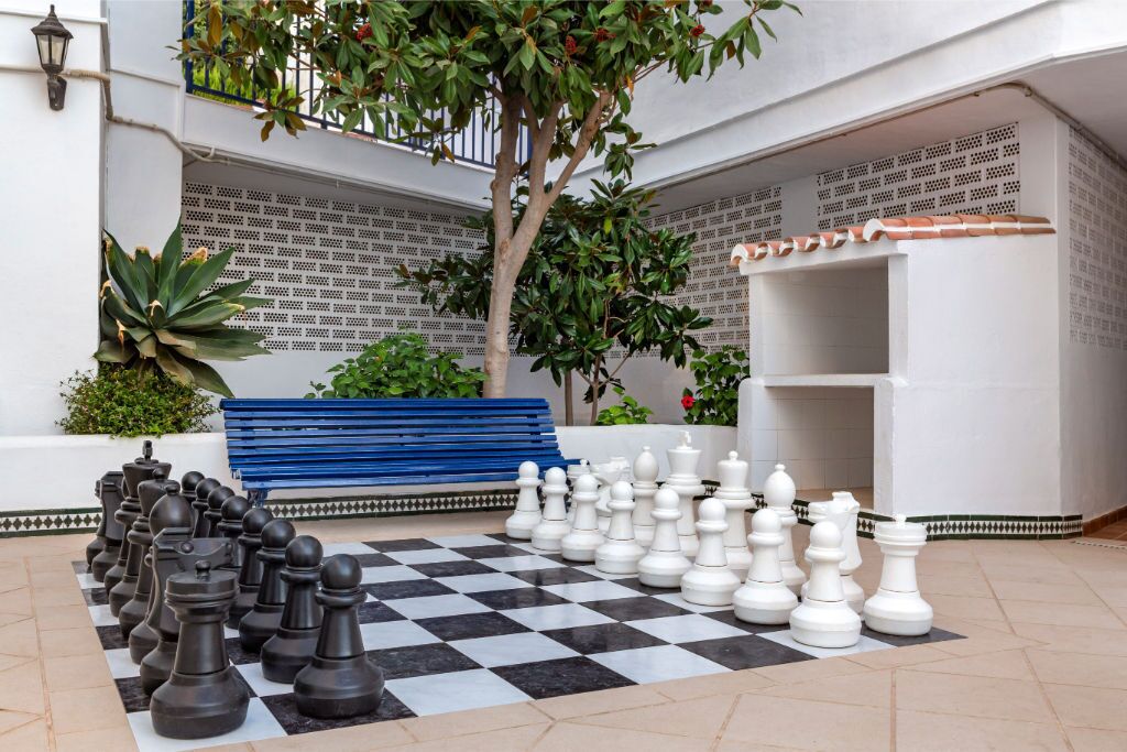 Ein Innenhof mit Schachbrett aus Schachfiguren, einer blauen Bank und einem Baum.