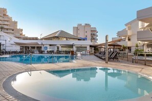 En swimmingpool med liggestole og et overdækket område, omgivet af moderne bygninger.