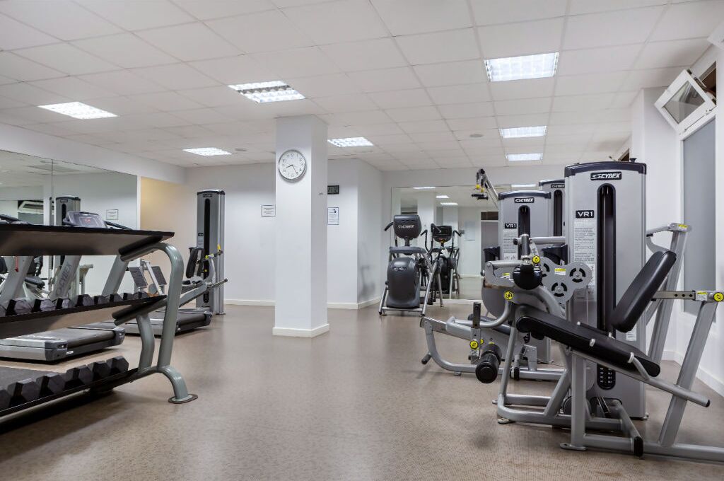 Uma academia com diversos equipamentos de exercício, um relógio na parede e um espelho refletindo os aparelhos.