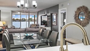 A modern living room with a glass-top dining table, grey upholstered seating, a chandelier, and a view of the water outside. - CLUB BAHIA WATERFRONT - COUNTRY CLUB USAGE (st . petersburg)