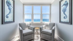 A room with two grey armchairs facing a window with a view of the ocean. There are two framed seahorse paintings on the walls.