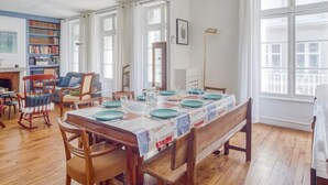 En matplats med ett träbord som är dukat för fyra personer, omgiven av trästolar. Till vänster finns en bokhylla med böcker och till höger ett stort fönster.