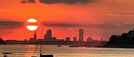 A sunset over a calm body of water with boats and a city skyline in the background.