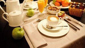 A breakfast table set with a coffee pot, cups, a plate of fruit, and a basket of pastries.