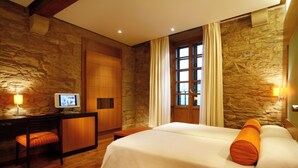 A hotel room with a stone wall, a bed with orange pillows, a desk with a computer, and a window with curtains.