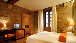 A hotel room with a stone wall, a bed with orange pillows, a desk with a computer, and a window with curtains. - Hotel Altair (Santiago de Compostela)