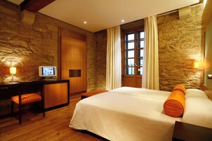 Una habitación de hotel con pared de piedra, una cama con almohadas anaranjadas, un escritorio con computadora y una ventana con cortinas.
