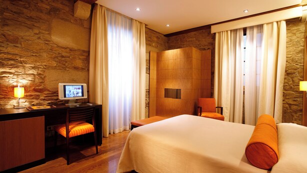 A hotel room with a stone wall, a wooden desk with a computer, a bed with orange pillows, and a shower area. - Hotel Altair (Santiago de Compostela)