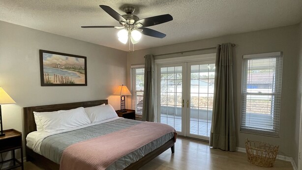 A bedroom with a bed, a ceiling fan, a nightstand, a lamp, a window with curtains, and a wicker basket.