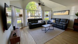 A spacious living room with a large window, a ceiling fan, a black leather sofa, a coffee table, and a flat-screen TV. - Fully Remodeled  stylish 3/2 condo, close to the parks (kissimmee)