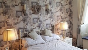 Un dormitorio con papel tapiz floral, una cama blanca con ropa de cama estampada con corazones, dos lámparas de noche y una mesita de noche con una lámpara y un pequeño adorno.