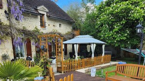 Un edificio de piedra con una terraza de madera, una cerca de madera y una pérgola con plantas colgantes.