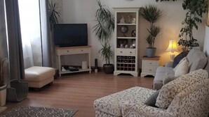 Una sala de estar con sofá, televisor sobre un mueble bajo, aparador con adornos y varias plantas en macetas.