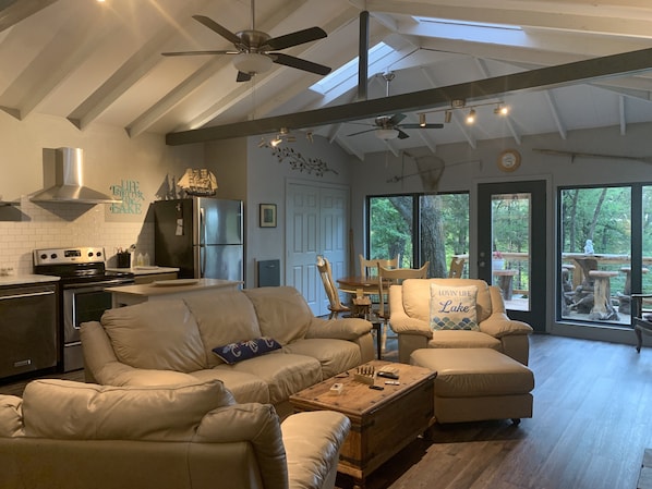 A spacious living room with a large sofa, a wooden coffee table, and a dining area with a table and chairs. There is a kitchen area with a stove, refrigerator, and ceiling fans. The room has large windows offering a view of the outdoors.
