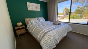A neatly made bed with patterned bedding, a small bedside table with a lamp, and a large window with a view of trees and buildings.