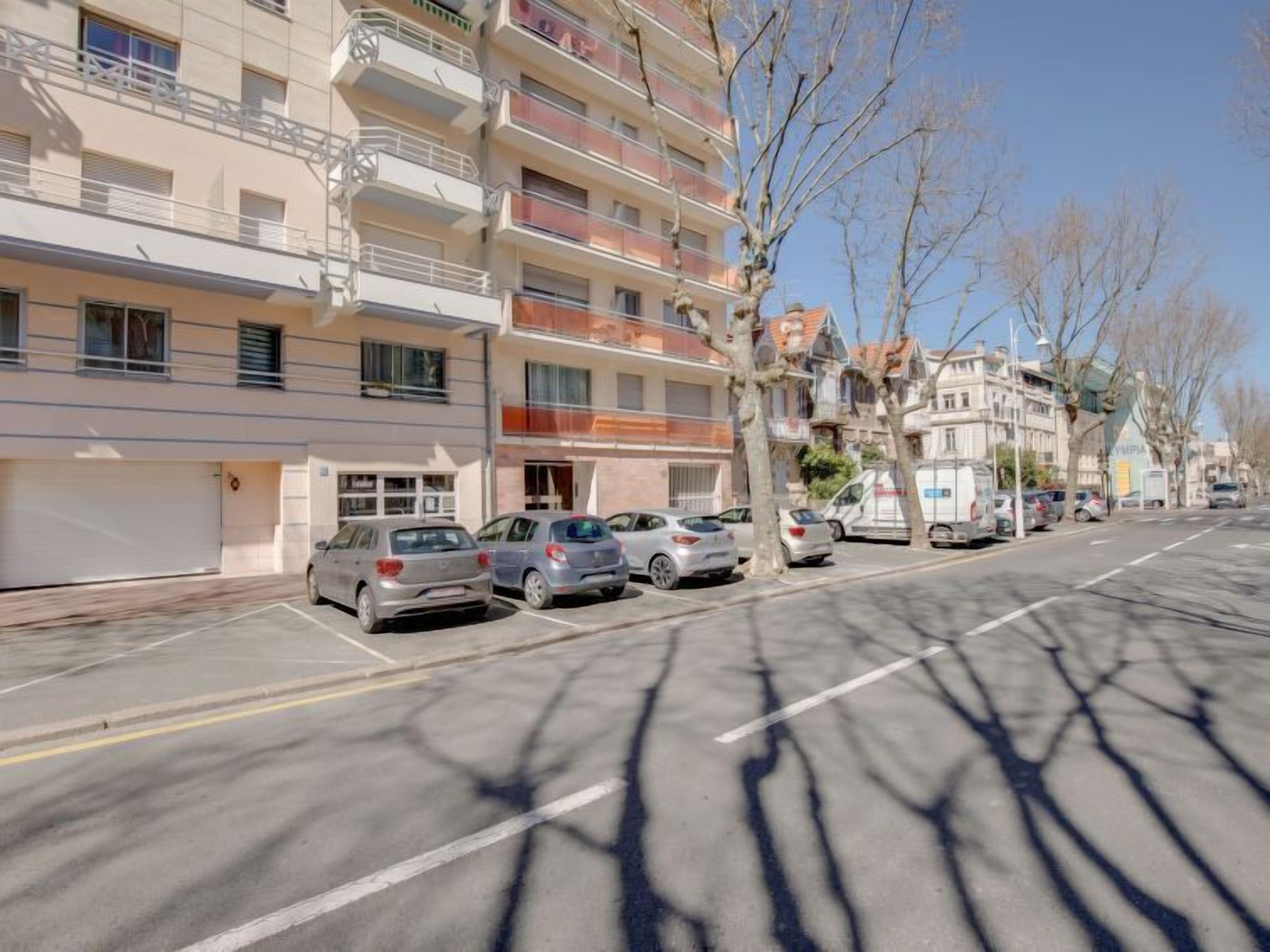 Un immeuble résidentiel à plusieurs étages, doté de balcons, avec des voitures garées et des arbres sans feuilles.