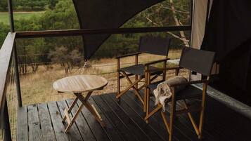 A wooden deck with a table and two chairs, a view of greenery through a window, and a dark canopy.