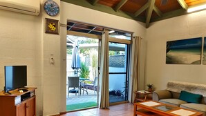 A living room with a TV, sofa, and coffee table, a sliding glass door to a patio, and a clock on the wall. - Beachside Cottage - A Private Peaceful Hidden Gem (Nelly Bay)