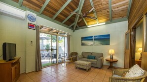 A living room with a television, a sofa, a chair, and a table. There is a ceiling fan and a painting on the wall. - Beachside Cottage - A Private Peaceful Hidden Gem (Nelly Bay)