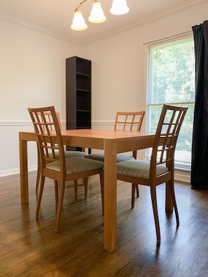 A dining area with a wooden table and chairs, a tall black bookshelf, and a window with blinds.