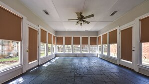 Une pièce spacieuse dotée d’un sol en pierre, de grandes fenêtres avec des stores bruns et d’un ventilateur de plafond.