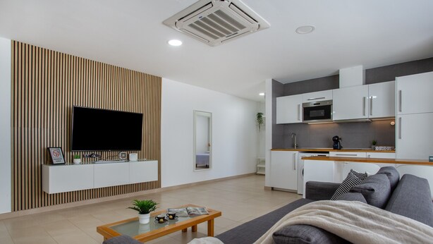 A modern living room with a flat-screen TV, a sofa, a coffee table, and a kitchen area with cabinets and appliances.