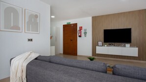 A modern living room with a grey sofa, a flat-screen TV mounted on a wooden panel wall, and a door labeled 'SALIDA' (exit).
