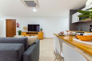Ein modernes Wohnzimmer mit einer Couch, einem Holzesstisch, einer Küchenzeile mit Mikrowelle und einem Fernseher.