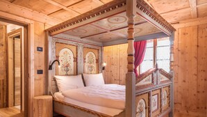 Un lit à baldaquin avec une tête de lit et un pied de lit richement décorés, un plancher en bois et une fenêtre ornée d’un rideau rouge.