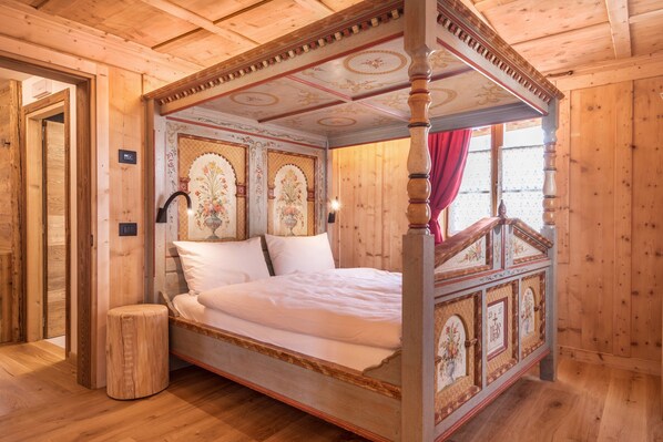 A four-poster bed with ornate headboard and footboard, a wooden floor, and a window with a red curtain.