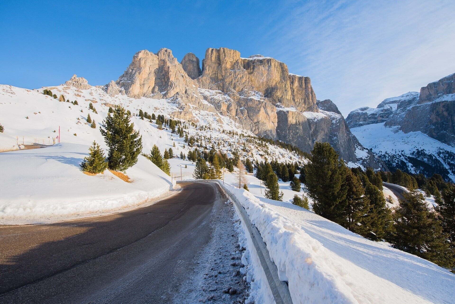 Une route de montagne enneigée, bordée d’une barrière de sécurité, d’arbres et dominée par un paysage montagneux rocheux.
