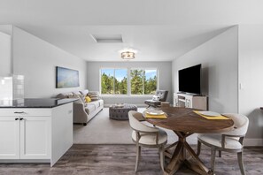 A modern kitchen with white cabinetry, a dining area with a wooden table and chairs, a gray ottoman, and a large window with a view of trees. - Dara 30 Day - Modern Irish Guest House on Private Estate, Mountain Views (Bend)