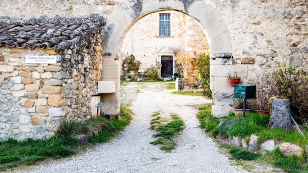 Une entrée du château Saint-Secret, avec un chemin de pierre menant à un bâtiment doté d’un toit en tuiles et d’une fenêtre.