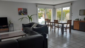 Ein modernes Wohnzimmer mit einer Couch, einem Esstisch mit Stühlen, einem Fernseher und einem großen Fenster mit Blick auf Grünflächen.