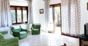 Una stanza con due poltrone verdi, un tavolino e una televisione. Sono presenti finestre con tende floreali e una porta che dà sull'esterno.