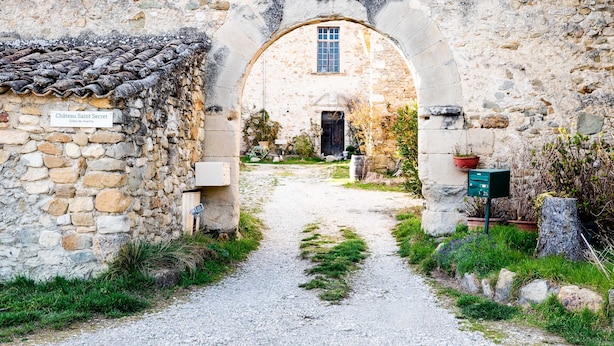 Una entrada al Château Saint Secret con un camino de piedra que lleva a un edificio con tejado de tejas y una ventana.
