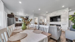 Eine moderne Küche mit Einbauofen, ein Essbereich mit Holztisch und weißen Stühlen sowie ein Wohnzimmer mit grauem Sofa und Wand-Fernseher.