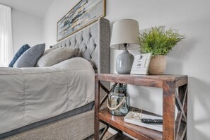 Un lit bien fait, avec une tête de lit grise, une lampe de chevet, une petite plante en pot et une table de chevet en bois sur laquelle se trouvent un bocal en verre et une télécommande.