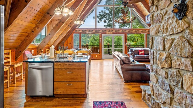 A spacious loft-style room with a large wooden ceiling, stone walls, a central island with a dishwasher, and a cozy seating area with a fireplace.