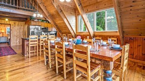 A spacious dining area with wooden tables and chairs, a kitchen in the background, and a large window providing a view of the outdoors.