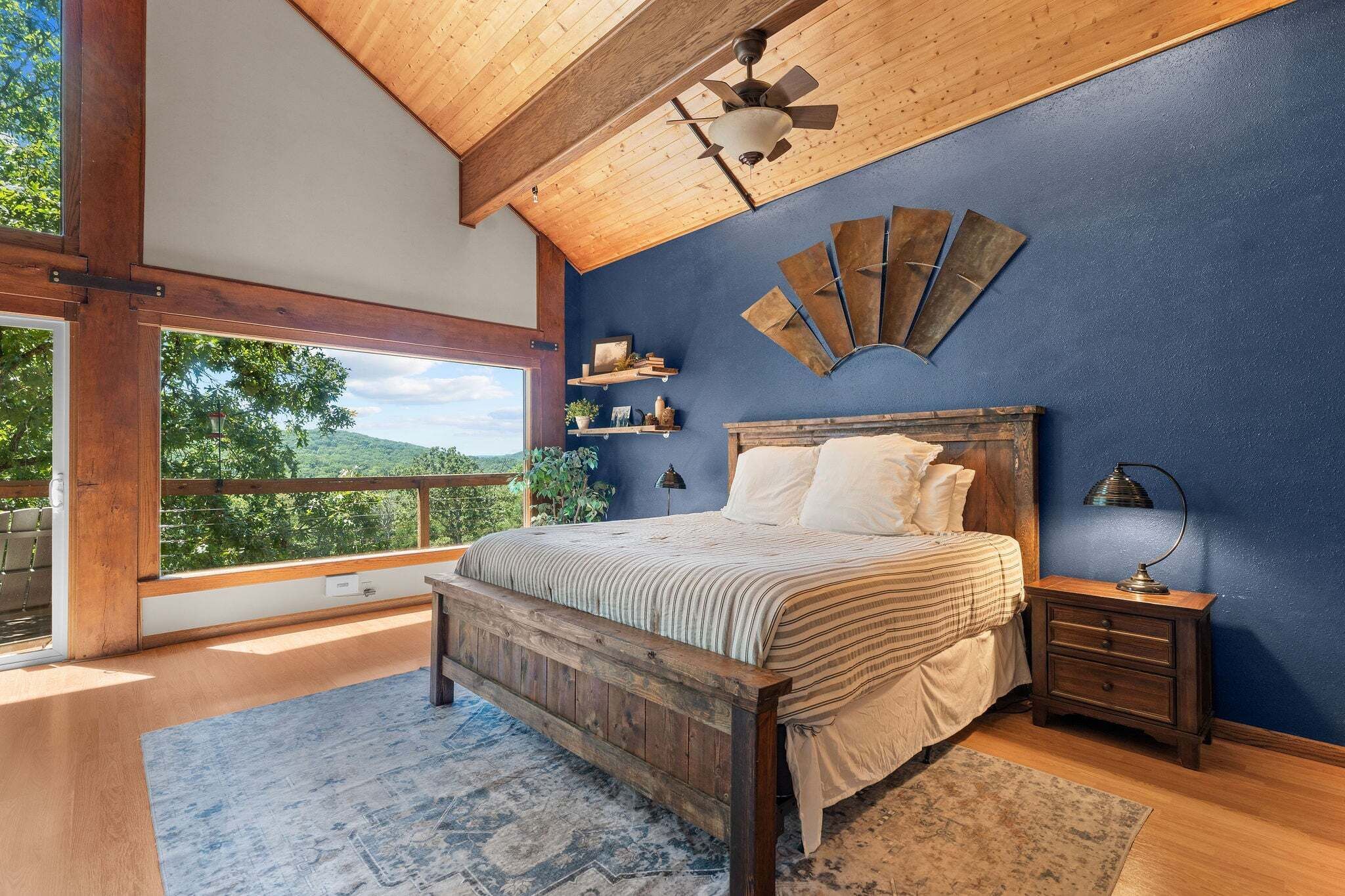 Une chambre avec un grand lit, des poutres apparentes en bois au plafond, une vue par la fenêtre et un mur d’accent bleu.