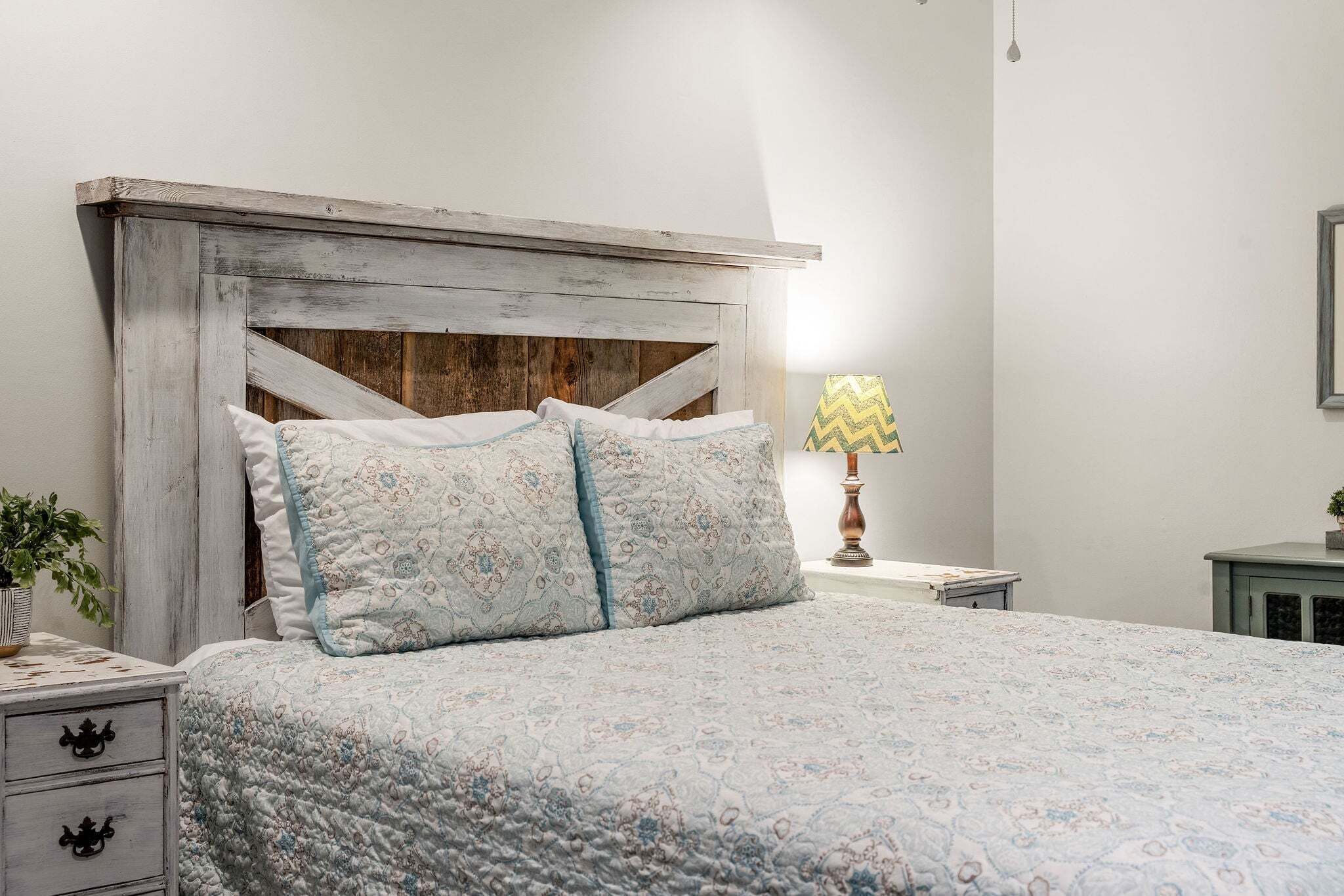Uma cama com cabeceira acolchoada, duas almofadas, uma luminária de cabeceira e uma mesa de cabeceira com gaveta.